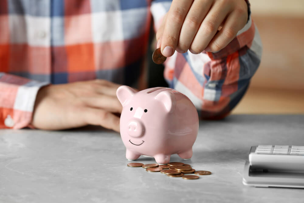 Man Putting Coin Into Piggy Bank at Grey Marble Table, Closeup. 