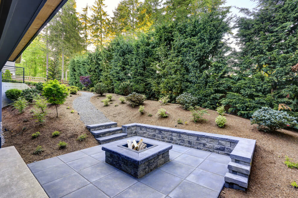 New Modern Home Features a Backyard With Rectangular Concrete Fire Pit Framed by Slate Pavers and Overlooking the Lush Garden.