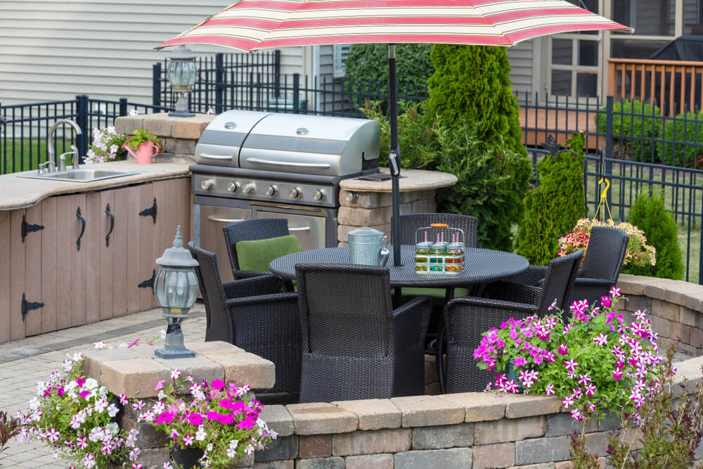 Outdoor kitchen on a brick exterior patio with wicker dining chairs under an umbrella.